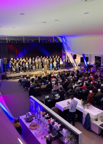 Das Bild zeigt einen Chor auf einer Bühne stehen. Vor der Bühne sind zahlreiche Stuhlreihen zu sehen. Beinahe alle Plätze sind gefüllt. Naben den Stuhlreihen sind zwei Tresen aufgebaut, an denen Getränke und Gläser stehen. Der Raum ist hell erleuchtet.    
