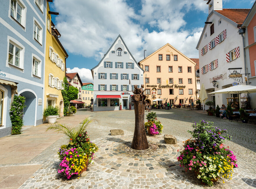 Das Bild zeigt eine gemütliche mittelalterliche Stadt mit farbenfrohen Gebäuden, Kopfsteinpflasterstraßen und Blumenkübeln. Die Stadt ist lebhaft mit Menschen befüllt und es gibt mehrere Sonnenschirme und Tische für eine gemütliche Einkehr.