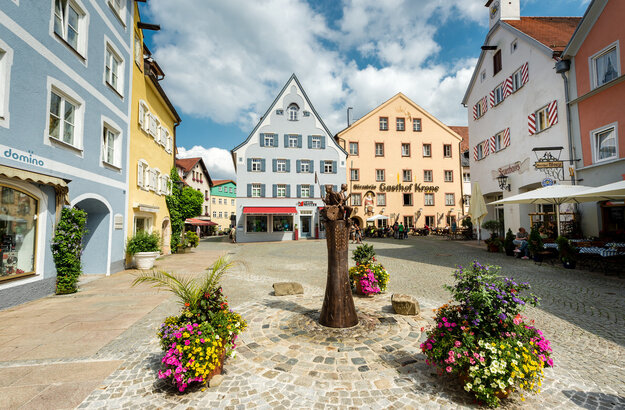 Das Bild zeigt eine gemütliche mittelalterliche Stadt mit farbenfrohen Gebäuden, Kopfsteinpflasterstraßen und Blumenkübeln. Die Stadt ist lebhaft mit Menschen befüllt und es gibt mehrere Sonnenschirme und Tische für eine gemütliche Einkehr.