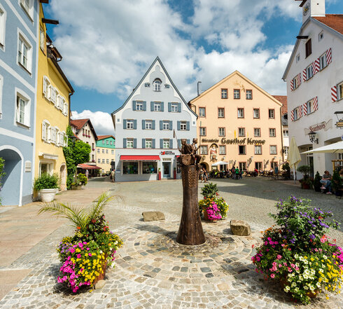 Das Bild zeigt eine gemütliche mittelalterliche Stadt mit farbenfrohen Gebäuden, Kopfsteinpflasterstraßen und Blumenkübeln. Die Stadt ist lebhaft mit Menschen befüllt und es gibt mehrere Sonnenschirme und Tische für eine gemütliche Einkehr.
