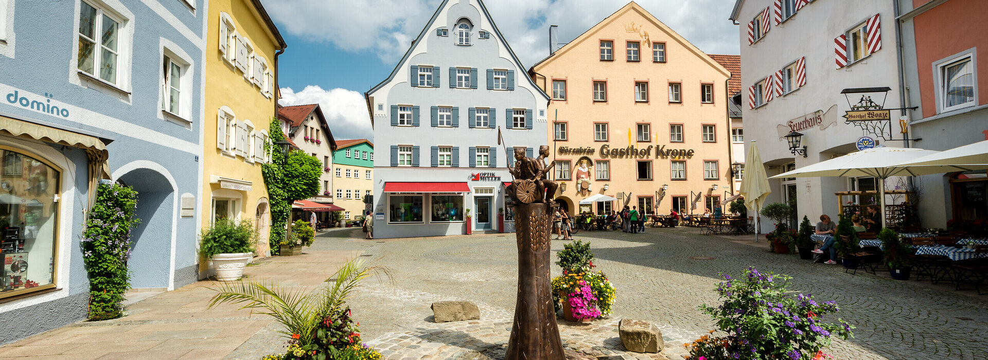 Das Bild zeigt eine gemütliche mittelalterliche Stadt mit farbenfrohen Gebäuden, Kopfsteinpflasterstraßen und Blumenkübeln. Die Stadt ist lebhaft mit Menschen befüllt und es gibt mehrere Sonnenschirme und Tische für eine gemütliche Einkehr.