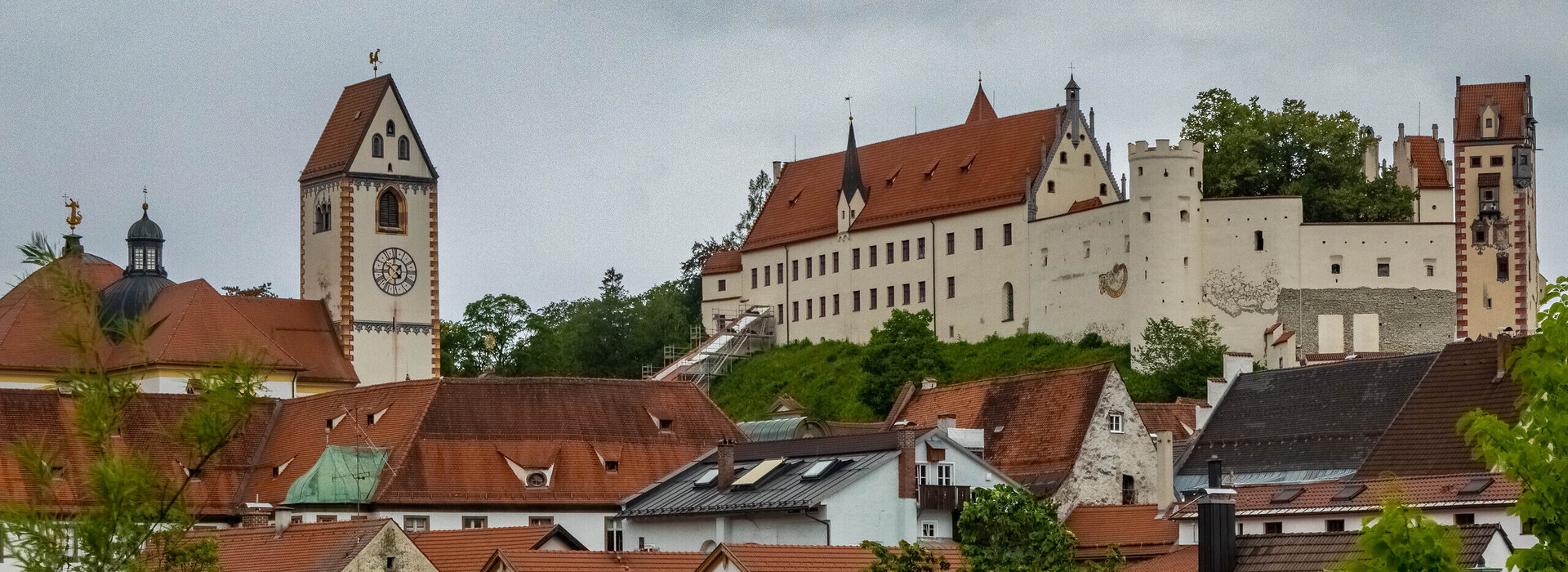 Das Foto zeigt das Hohe Schloss und die Kirche St. Mang über den Dächern von Füssen. Der Himmel ist grau und wolkenverhangen.  