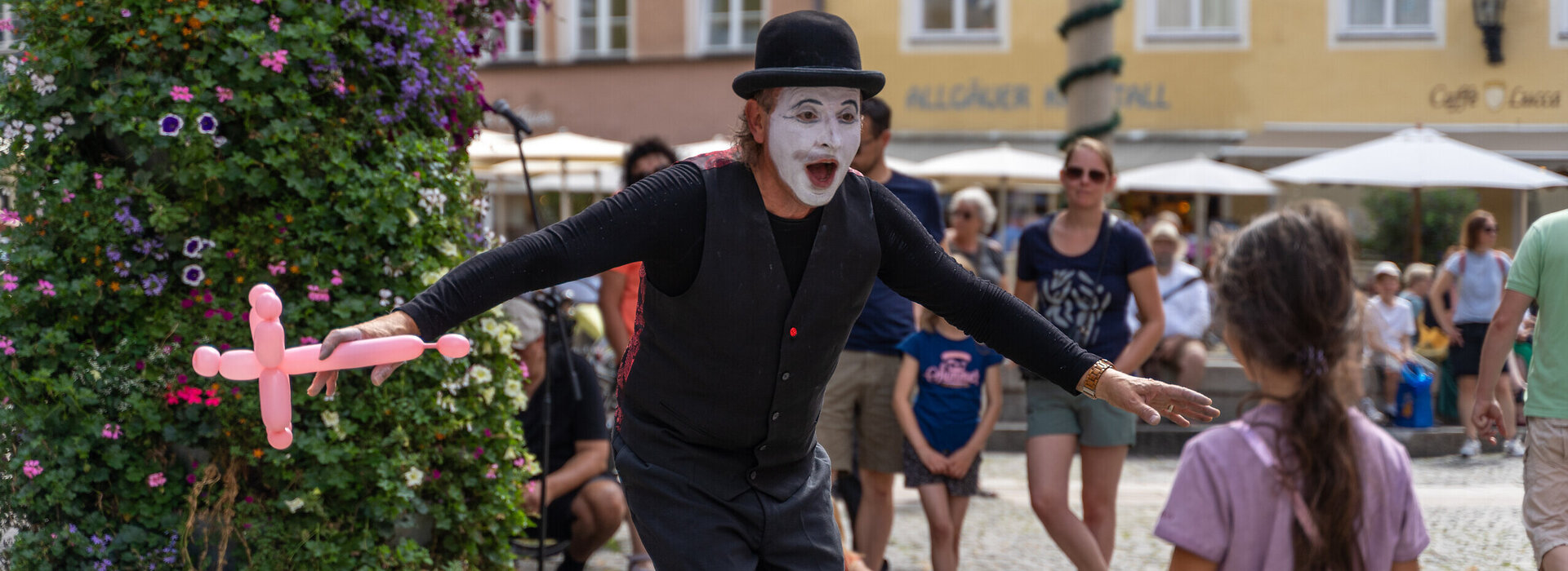 Das Bild zeigt einen Pantomimen, der ein Luftballontier in der rechten Hand hält. Er ist schwarz gekleidet, trägt einen schwarzen Hut und ist weiß geschminkt. Er steht in einer Fußgängerzone. Mehrere Personen sind zu sehen. Im Hintergrund sieht man bunte Hausfassaden. Die Sonne scheint. 