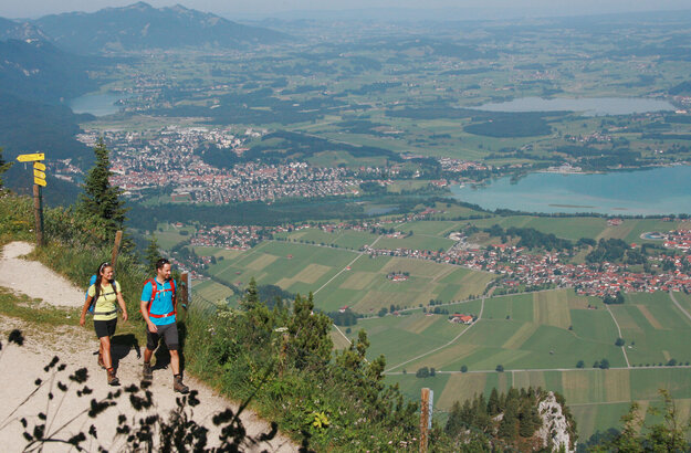Ein Paar wandert auf dem Gipfel des Tegelbergs. Im Hintergrund sind Füssen, der Forggensee und die Tannheimer Alpen zu sehen