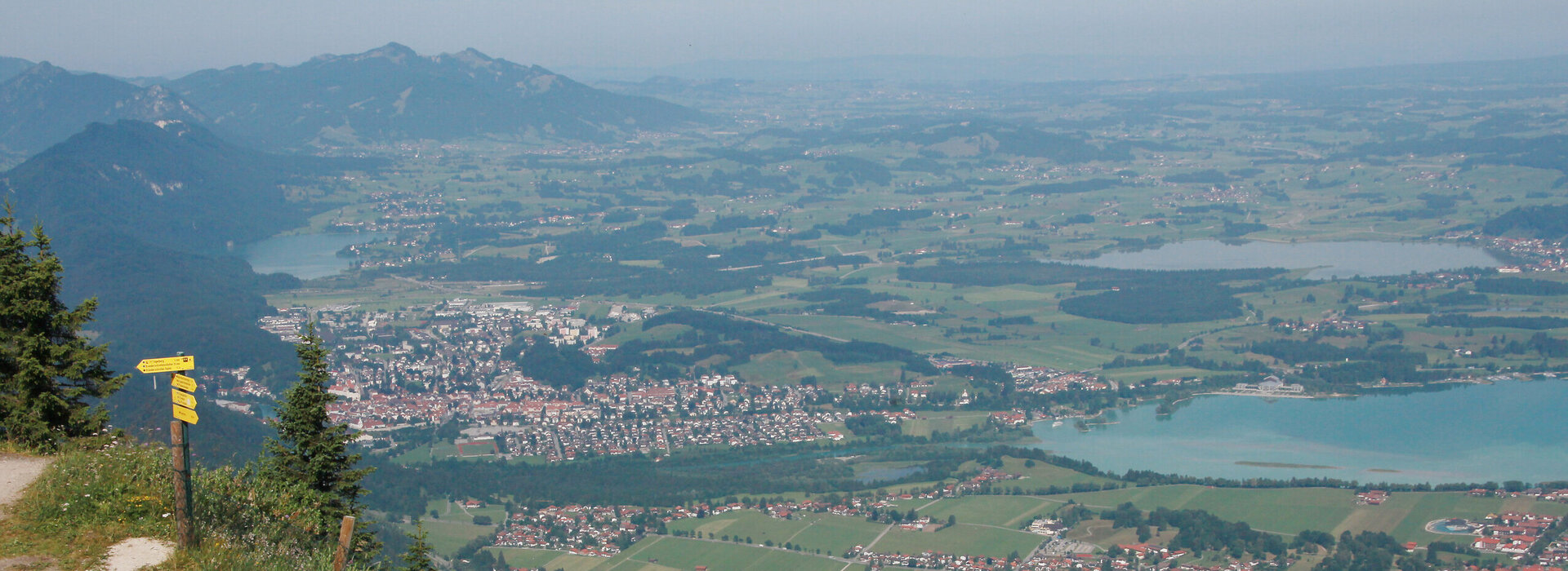 Ein Paar wandert auf dem Gipfel des Tegelbergs. Im Hintergrund sind Füssen, der Forggensee und die Tannheimer Alpen zu sehen