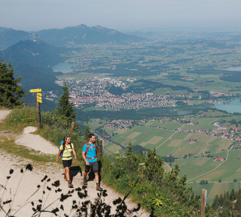 Ein Paar wandert auf dem Gipfel des Tegelbergs. Im Hintergrund sind Füssen, der Forggensee und die Tannheimer Alpen zu sehen