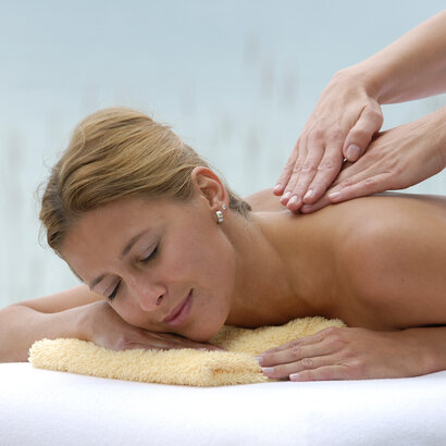 Zu sehen ist eine junge Frau, die oberkörperfrei in Bauchlage auf einer Liege liegt. Ihr Kopf ist auf ein gelbes, zusammengelegtes Handtuch gebettet. Sie lächelt. Vom rechten Bildrand kommend sind zwei weitere Hände zu sehen, die der Frau den Rücken massieren.