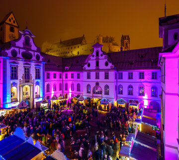 Das Bild fängt eine lebendige nächtliche Szene in Füssen auf dem Adventsmarkt ein. Die Skyline wird dominiert von einem großen, verzierten Gebäude mit Uhrturm, das sich deutlich gegen den Nachthimmel abhebt. Das Gebäude ist mit lila und pinken Lichtern beleuchtet, was eine fröhliche Atmosphäre schafft. Die Straße vor dem Gebäude ist von Menschen belebt, die ebenfalls von den gleichen lila und pinken Lichtern erhellt werden. Die Menge scheint ein Fest oder eine Veranstaltung zu genießen, wie die bunten Lichter und die lebendige Atmosphäre darauf hindeuten. Die Gesamtansicht vermittelt das Gefühl von Feier und Gemeinschaftsgeist.