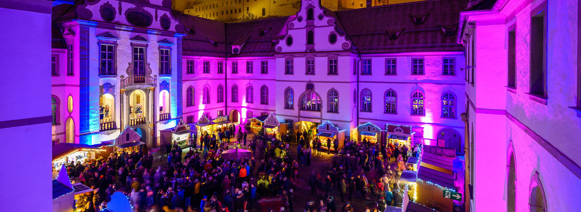 Das Bild fängt eine lebendige nächtliche Szene in Füssen auf dem Adventsmarkt ein. Die Skyline wird dominiert von einem großen, verzierten Gebäude mit Uhrturm, das sich deutlich gegen den Nachthimmel abhebt. Das Gebäude ist mit lila und pinken Lichtern beleuchtet, was eine fröhliche Atmosphäre schafft. Die Straße vor dem Gebäude ist von Menschen belebt, die ebenfalls von den gleichen lila und pinken Lichtern erhellt werden. Die Menge scheint ein Fest oder eine Veranstaltung zu genießen, wie die bunten Lichter und die lebendige Atmosphäre darauf hindeuten. Die Gesamtansicht vermittelt das Gefühl von Feier und Gemeinschaftsgeist.