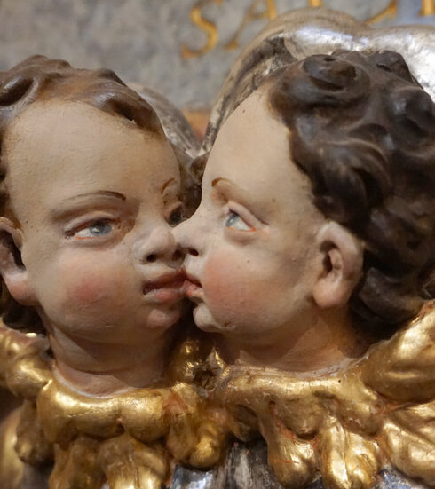 Das Foto zeigt die Köpfe zweier Putten, die sich an Mund und Nase berühren. Sich also scheinbar küssen. Die Puttenköpfe haben braune Haare und blaue Augen. An der Stelle der Kragen sind goldene Engelsflügel zu sehen. 