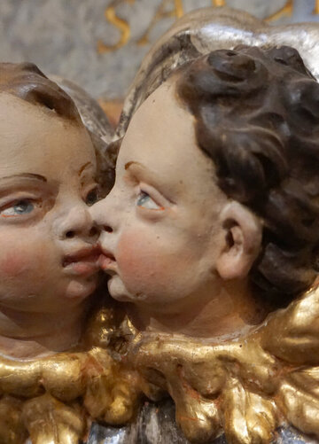 Das Foto zeigt die Köpfe zweier Putten, die sich an Mund und Nase berühren. Sich also scheinbar küssen. Die Puttenköpfe haben braune Haare und blaue Augen. An der Stelle der Kragen sind goldene Engelsflügel zu sehen. 