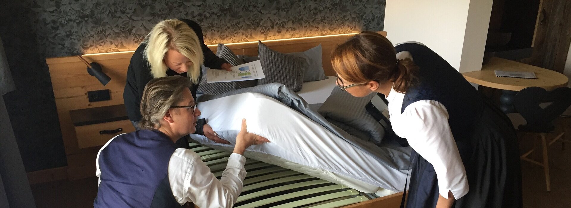 Das Bild zeigt drei Personen, die die Matratze eine Bettes hochheben und den darunterliegenden Lattenrost betrachten. Eine Frau trägt ein dunkles Dirndl. Eine Frau hat Papiere in der Hand. Ein Mann in Jeans, weißem Hemd und Weste ist in die Hocke gegangen.