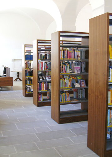 Man blickt in einen hohen, hellen Raum mit Kreuzrippengewölbe. In der Mitte des Raumes stehen hohe, hölzerne Bücherregale voller Bücher. Der Boden ist mit steinernen Fließen ausgelegt. Im Hintergrund steht ein Schreibtisch mit einem Computer darauf.