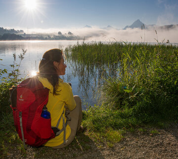 Das Bild zeigt eine Frau in Wanderkleidung, die am Ufer eines Sees sitzt. Sie trägt einen roten Rucksack. Über dem See sind Nebelschwanden zu sehen. Ufernah ist ein Schildgürtel zu sehen. Im Hintergrund erheben sich hohe Berge. Der Himmel ist blau. Die Sonne scheint.     