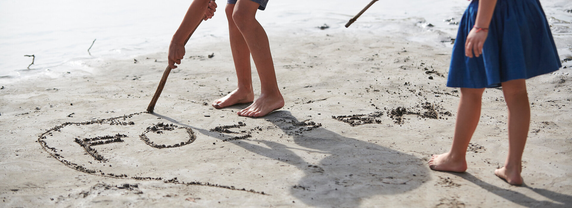 Das Bild zeigt eine ruhige Strandszene mit zwei jungen Kindern, die sich im Vordergrund befinden. Das Kind auf der linken Seite, das ein gestreiftes Hemd mit blauen und weißen Streifen und blaue Shorts trägt, beugt sich über die Erde, um einen Stock aufzuheben. Das Kind auf der rechten Seite, das ein rosa Shirt und einen blauen Rock trägt, hält einen Stock und weist damit auf das Wasser. Der sandige Strand ist mit kleinen Steinen und Kieseln gesprenkelt, was der Szene Textur verleiht. Das ruhige Wasser im Hintergrund vervollständigt die idyllische Kulisse.