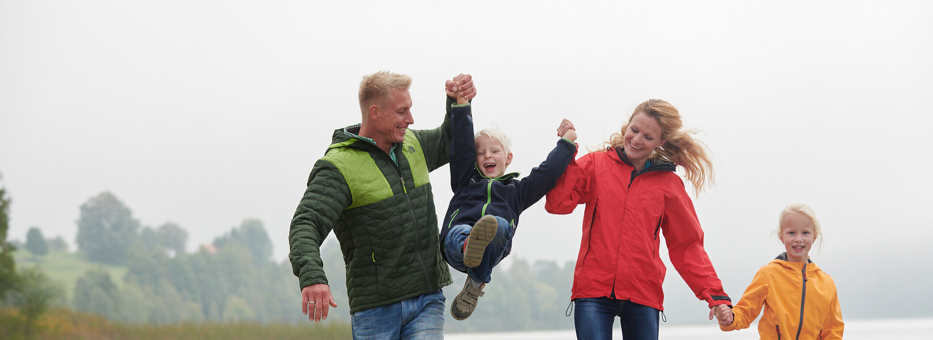 Das Bild fängt eine herzerwärmende Szene einer vierköpfigen Familie ein, die gemeinsam entlang eines sandigen Strandes geht. Der Vater trägt ein grünes Jackett und hält die Hände seiner zwei Kinder, die jeweils ein gelbes Jackett tragen. Die Mutter trägt ein rotes Jackett und hält die Hand des Vaters. Sie alle gehen Richtung Wasser, das im Hintergrund sichtbar ist. Die Familie scheint einen schönen Tag am Strand zu genießen. Der sandige Strand erstreckt sich im Vordergrund, während das Wasser eine beruhigende Kulisse für die Szene schafft. Die Garderobe der Familie deutet darauf hin, dass es möglicherweise ein kühler Tag ist und sie angemessen für das Wetter gekleidet sind. Das Bild vermittelt ein Gefühl von Zusammengehörigkeit und Naturgenuss.