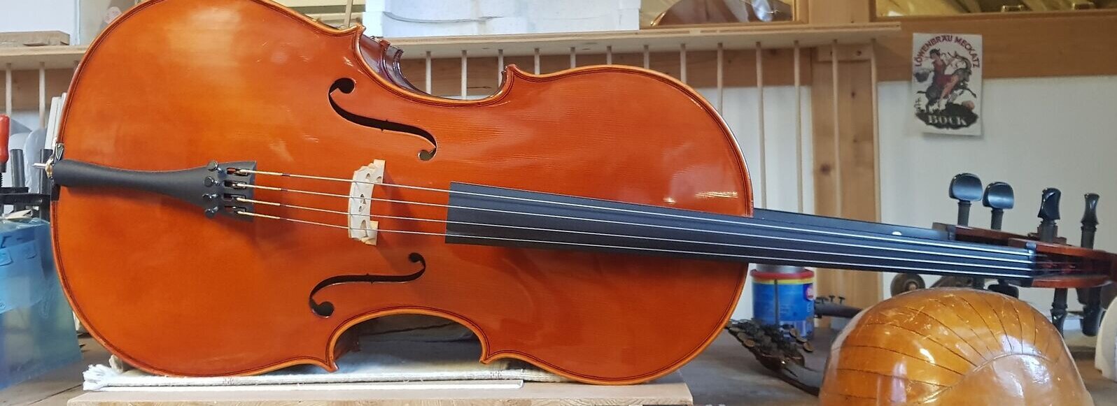 Das Foto zeigt ein Cello, das auf der Seite liegt. Unter dem Celloarm liegt der Corpus einer Laute. Im Hintergrund sind unterschiedliche Hölzer und Werkzeuge zu sehen. Der Raum ist hell und einladend. 