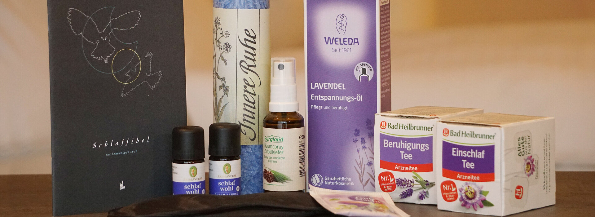 Das Bild zeigt unterschiedliche Schlafgoodies. Von links nach rechts: Ein dunkelgraues Heft mit der Aufschrift „Schlaffibel“. Zwei kleine braune Fläschchen mit der Aufschrift „schlaf wohl“, eine große hellblaue Kerze mit der Aufschrift „Innere Ruhe“, kleine braune Sprayflasche mit der Aufschrift „Bergland, Raumspray Zirbelkiefer“, eine hohe, lila Schachtel mit der Aufschrift „Weleda, Lavendel, Entspannungs-Öl“, zwei kleine Schachteln mit je der Aufschrift „Bad Heilbrunner Beruhigungs-Tee, Arzneitee“, eine schwarze Schlafmaske und einen Teebeutel.