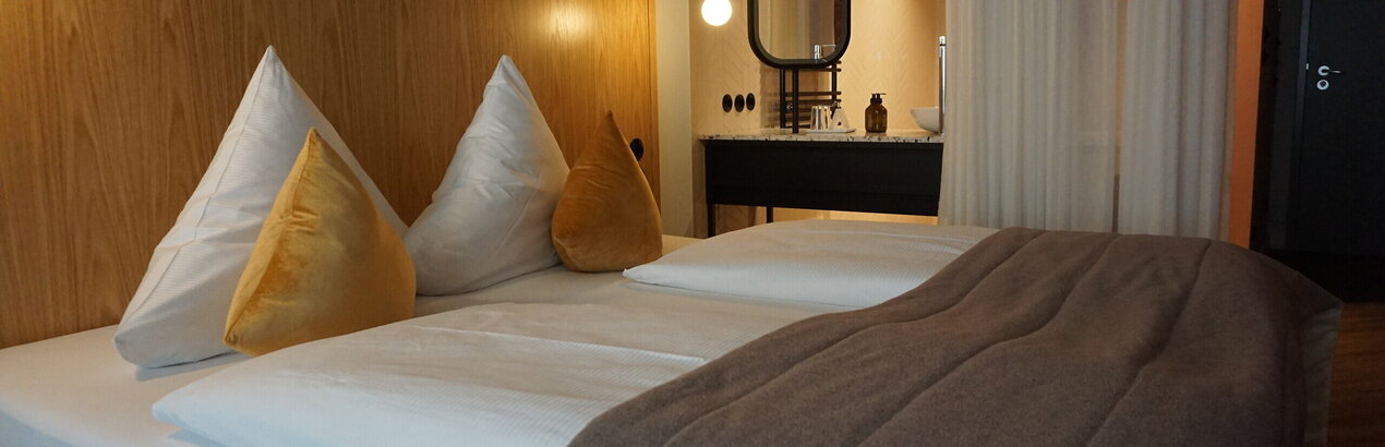  Das Bild zeigt ein modernes Hotelzimmer, das ein Gefühl von Luxus und Komfort vermittelt. Dominierend ist das große Bett, das in weiße Leintücher gehüllt ist und mit zwei goldfarbenen Kissen geschmückt ist. Das Bett steht an einer hölzernen Wand, die dem Raum eine warme Note verleiht.