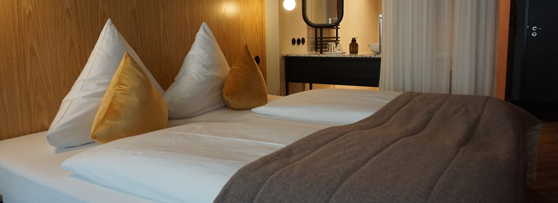  Das Bild zeigt ein modernes Hotelzimmer, das ein Gefühl von Luxus und Komfort vermittelt. Dominierend ist das große Bett, das in weiße Leintücher gehüllt ist und mit zwei goldfarbenen Kissen geschmückt ist. Das Bett steht an einer hölzernen Wand, die dem Raum eine warme Note verleiht.