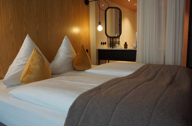  Das Bild zeigt ein modernes Hotelzimmer, das ein Gefühl von Luxus und Komfort vermittelt. Dominierend ist das große Bett, das in weiße Leintücher gehüllt ist und mit zwei goldfarbenen Kissen geschmückt ist. Das Bett steht an einer hölzernen Wand, die dem Raum eine warme Note verleiht.