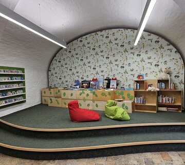 Auf dem Bild ist der Innenraum eines Vorleseraumes zu sehen. Es gibt mehrere Regale mit Büchern gefüllt und einen grünen und einen roten Sitzsack.
