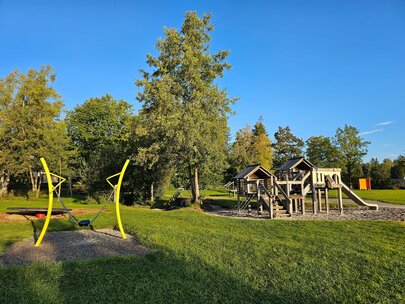 Auf dem Bild sieht man einen Spielplatz mit mehreren Spielgeräten. Im Vordergrund befindet sich eine grüne Schaukel. Etwas dahinter eine Drehscheibe und ein Rutschehaus. Alles ist von Bäumen umgeben. Es ist sonniges Wetter mti wolkenlosem Himmel. 