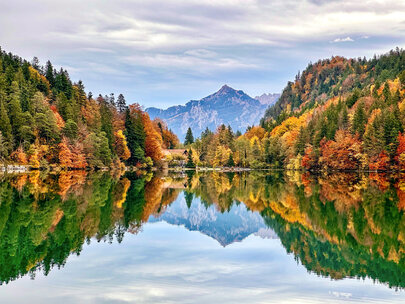 Im Fokus des Bildes liegt ein See, dessen Oberfläche glatt wie ein Spiegel, die umliegende Natur widerspiegelt. Den See säumen bunte Wälder. Im Hintergrund erheben sich die Berge.  