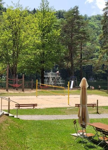 Das Bild zeigt einen Blick ins Gelände des Oberseebads. Das zentrale Motiv ist ein sandiger Volleyballplatz, umgeben von üppigem Grün, Bäumen und Gras. Der Platz ist mit weißen Linien markiert, die die Grenzen für das Spiel anzeigen. Im Vordergrund befinden sich mehrere hölzerne Bänke und Tische, die in einem Halbkreis um den Platz angeordnet sind.  In der Ferne liegt der ruhige Obersee. Die Oberfläche des Wassers ist unberührt und spiegelt die umliegende Vegetation wider. Der Himmel darüber ist klar und blau mit einzelnen, verstreuten Wolken.