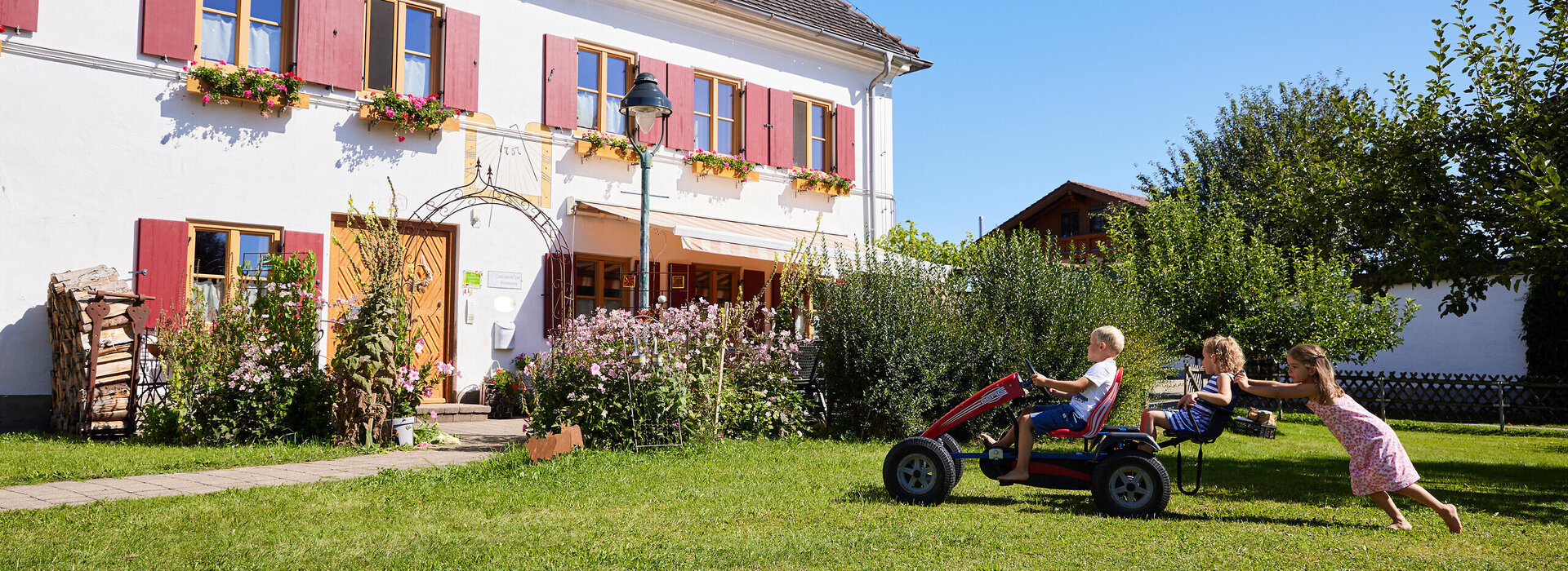 Das Bild zeigt eine lebhafte Szene in einem Wohngebiet. In der Mitte des Bildes ist eine Gruppe von Kindern zu sehen, die eine Fahrt auf einem kleinen, roten Dreirad genießen. Das Dreirad wird von einem Mädchen gezogen, das neben ihm läuft. Die Kinder auf dem Dreirad scheinen eine tolle Zeit zu haben, wobei eines sogar das Haar der Läuferin festhält, während sie fahren.  Das Dreirad steht vor einem großen, weißen Haus mit rotem Dach. Das Haus hat mehrere Fenster und einen kleinen Garten vor ihm. Der Garten ist mit pinken Blumen geschmückt und fügt der Szene einen Hauch von Farbe hinzu.  Im Hintergrund sind andere Häuser zu sehen, was darauf hindeutet, dass es sich um ein Wohngebiet handelt. Der Himmel über ihnen ist klar und blau, was auf einen sonnigen Tag hindeutet. Die Gesamtatmosphäre des Bildes ist eine von Fröhlichkeit und Spielfreude.