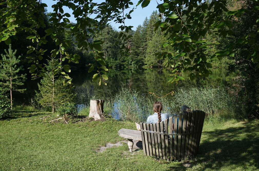 Das Bild zeigt im Vordergrund eine hölzerne Bank mit Rückenlehne, die sich auf einer grünen Wiese befindet. Eine Frau sitzt mit dem Rücken zur Kamera auf der Bank, ihre Haare sind zu einem Zopf gebunden. Sie scheint auf den vor ihr liegenden See, der von Bäumen und Vegetation umgeben ist, zu blicken. Das Wasser reflektiert die Vegetation und schafft eine beruhigende Atmosphäre. Der Himmel ist blau. 