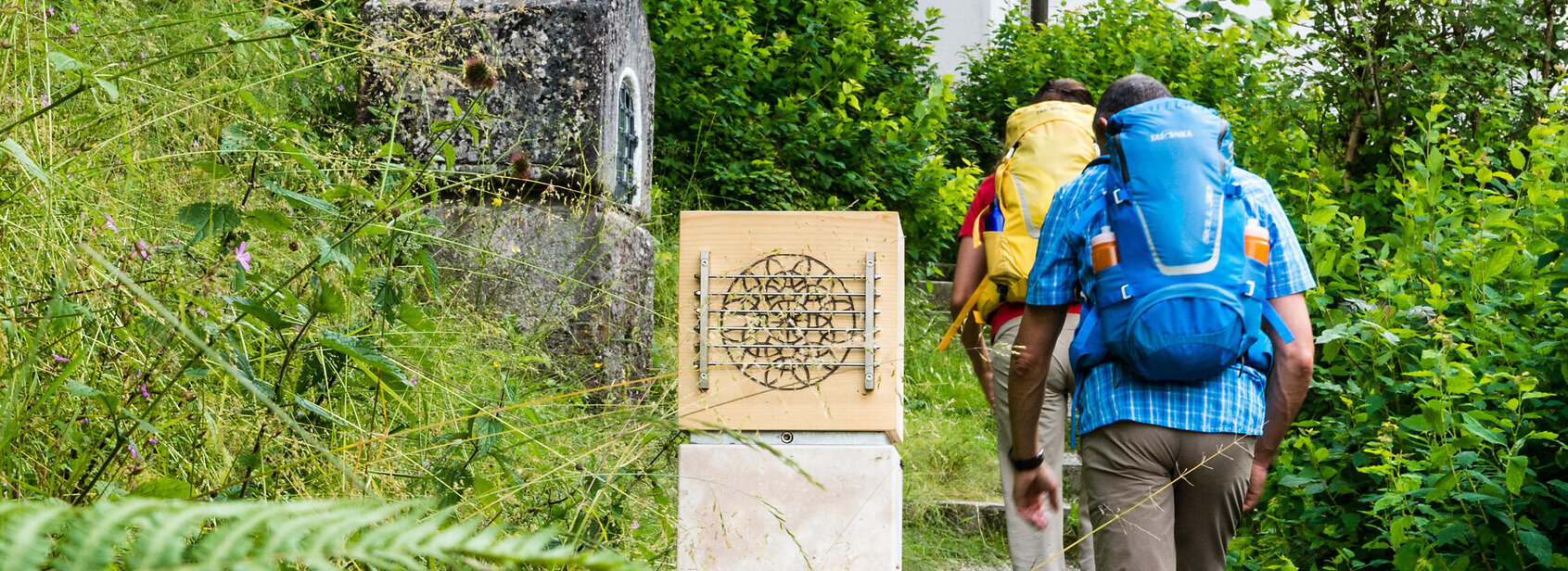 Das Bild zeigt einen Mann und eine Frau in Wanderkleidung mit Rucksäcken. Sie wandern einen Hang empor auf einem Kiesweg mit Stufen. Im Vordergrund steht eine steinerne Säule. Um sie herum wachsen Büsche, Bäume und Gräser.   