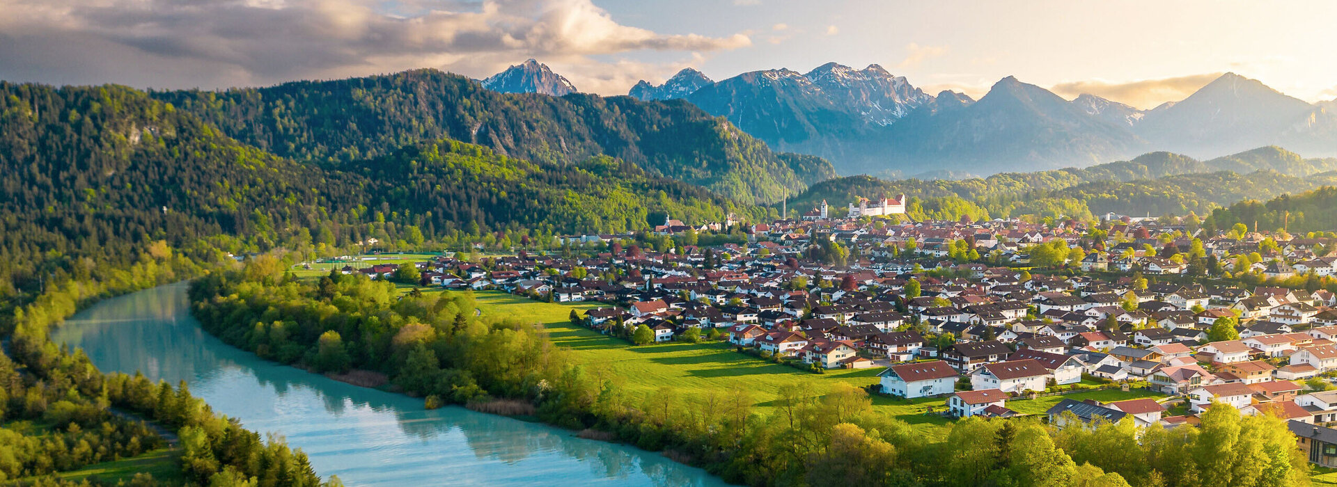 Das Bild zeigt einen breiten türkisblauen Fluss, der ruhig vor sich hinfließt. Am Ufer des Flusses wachsen Bäume. Auf der linken Uferseite beginnt der Bergwald. Am rechten Ufer erstreckt sich eine Wiese, die an die Stadt Füssen grenzt. Über Füssen thront auf einer Anhöhe das Hohe Schloss. Im Hintergrund ragen, teilweise schneebedeckte Berge empor. Die die Stadt und den Fluss einrahmen.  