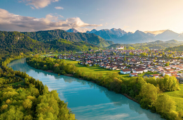 Das Bild zeigt einen breiten türkisblauen Fluss, der ruhig vor sich hinfließt. Am Ufer des Flusses wachsen Bäume. Auf der linken Uferseite beginnt der Bergwald. Am rechten Ufer erstreckt sich eine Wiese, die an die Stadt Füssen grenzt. Über Füssen thront auf einer Anhöhe das Hohe Schloss. Im Hintergrund ragen, teilweise schneebedeckte Berge empor. Die die Stadt und den Fluss einrahmen.  