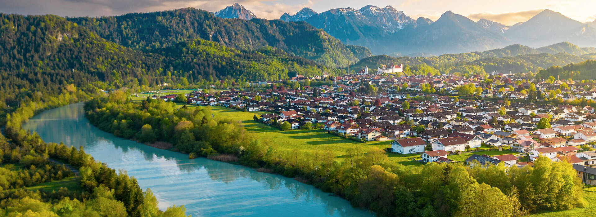 Das Bild zeigt einen breiten türkisblauen Fluss, der ruhig vor sich hinfließt. Am Ufer des Flusses wachsen Bäume. Auf der linken Uferseite beginnt der Bergwald. Am rechten Ufer erstreckt sich eine Wiese, die an die Stadt Füssen grenzt. Über Füssen thront auf einer Anhöhe das Hohe Schloss. Im Hintergrund ragen, teilweise schneebedeckte Berge empor. Die die Stadt und den Fluss einrahmen.  