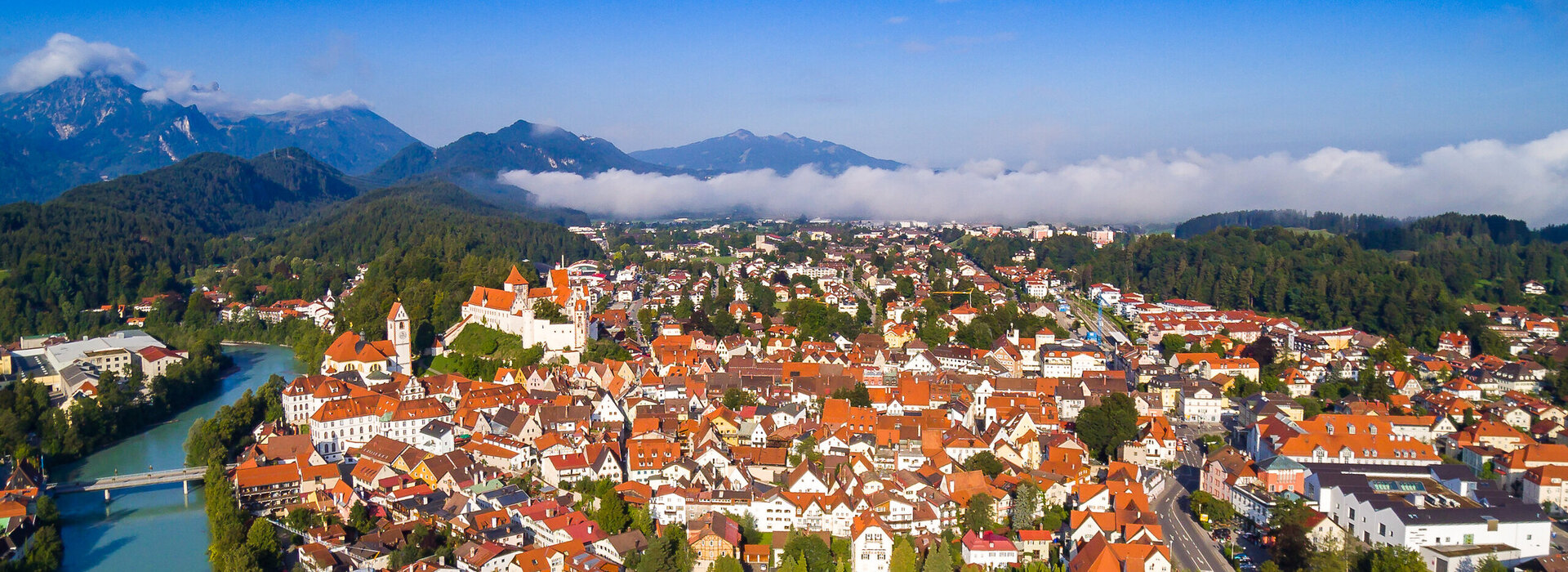 Das Foto zeigt Füssen von oben. Den Blick des Betrachters wird über die Dächer von Füssen geleitet. Im Vordergrund ist das Franziskanerkloster zu erkennen, das aus Klostergebäude und Klosterkirche St. Stephan besteht. Das Klostergelände ist eingerahmt von der historischen Stadtmauer. Neben dem Kloster fließt der türkisblau Fluss Lech. Im Hintergrund des Klosters erstreckt sich die Füssener Altstadt mit dem Hohen Schloss und dem Bergpanorama.