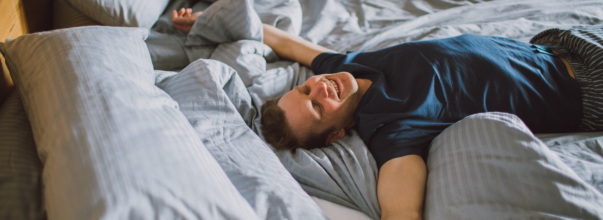 Das Bild zeigt einen Mann in einem kurzen Pyjama. Er sitzt liegt auf einem Doppelbett. Hat die Hände und Arme von sich gestreckt, die Augen geschlossen und lächelt. 
