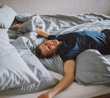 Das Bild zeigt einen Mann in einem kurzen Pyjama. Er sitzt liegt auf einem Doppelbett. Hat die Hände und Arme von sich gestreckt, die Augen geschlossen und lächelt. 