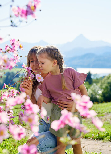 Die Frau und das Kind stehen in einem Garten, der voll von blühenden Pflanzen ist. Die Frau hält einen Strauß Blumen in der Hand und schneidet eine Blume. Das Kind, das auf dem Rücken der Frau sitzt, schaut in die Blumen. Beide sind in einem Moment der Natur zugewandt und genießen die Schönheit der Blumen.  Der Garten liegt in einer hügeligen Landschaft, die von einer Bergkette umgeben ist. Die Blumen sind in verschiedenen Farben, darunter auch in Pink, was die Atmosphäre des Gartens beeinflusst.  Die Frau und das Kind sind in einem Moment der Natur zugewandt und genießen die Schönheit der Blumen. Der Garten liegt in einer hügeligen Landschaft, die von einer Bergkette umgeben ist. Die Blumen sind in verschiedenen Farben, darunter auch in Pink, was die Atmosphäre des Gartens beeinflusst.