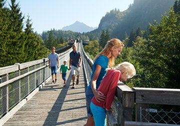 Das Bild fängt eine friedliche Szene auf einer Holzbrücke ein. Ein Familienvater ist mit einer blauen Jacke als dominantes Motiv abgebildet und führt die Familie über die Brücke. Seine Frau mit einer rosafarbenen Jacke folgt ihm. Ihre beiden Kinder, eine in einer grünen Jacke und die andere in einer roten Jacke, laufen dicht hinter ihnen. Die Brücke, die sie betreten haben, ist aus Holz und wird von üppig grünen Bäumen und einem klaren blauen Himmel umgeben. In der Ferne sind majestätische Berge zu sehen, die zur Schönheit der Szene beitragen. Die Familie scheint die Zeit miteinander zu genießen und erfreut sich am beeindruckenden Blick auf die Berge.