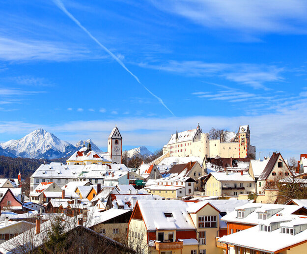 Das Foto zeigt die verschneiten Dächer von Füssen. Über den Dächern thront auf deiner Anhöhe das Hohe Schloss, daneben die St. Mang Klosterkirche. Im Hintergrund sind die verschneiten Berge zu sehen. Der Himmel ist blau. Die Sonne scheint. Es ist Winter.