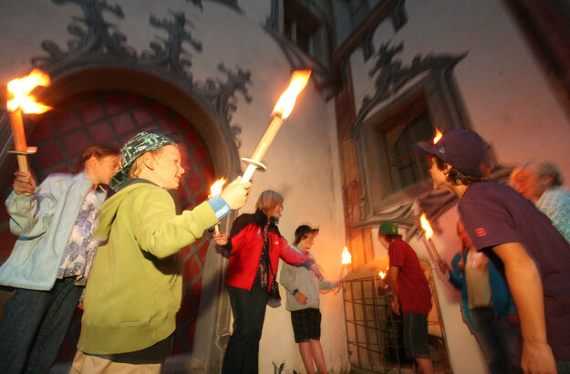 Das Bild zeigt eine lebendige Szene einer Gruppe von Menschen, die sich vor einem Gebäude versammeln. Das Gebäude, gekennzeichnet durch einen großen Bogen, dient als Kulisse für die Gruppe. Die Menschen, ein Gemisch aus Erwachsenen und Kindern, sind mit dem Aktes des Kerzenanzündens beschäftigt. Die Kerzen, von der Gruppe erhoben, brennen, und werfen ein warmes Schein auf die Szene. Die Gruppe ist vielfältig, mit Individuen unterschiedlichen Alters und Geschlechts. Einige von ihnen halten Fackeln, was zur feierlichen Stimmung beiträgt. Das Gesamtbild ist eine Feier und Einheit.