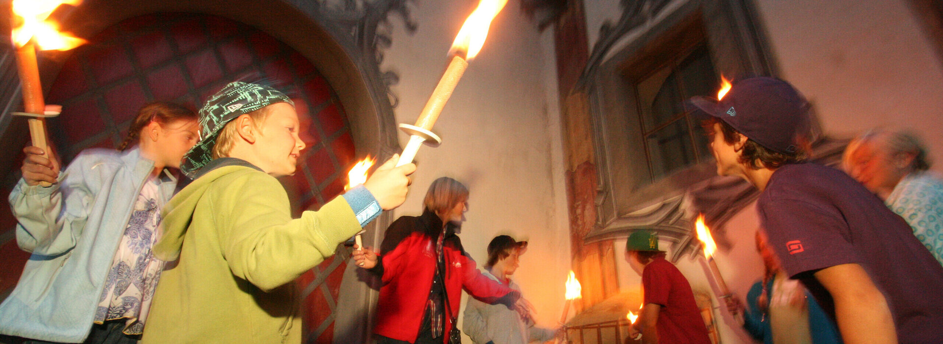 Das Bild zeigt eine lebendige Szene einer Gruppe von Menschen, die sich vor einem Gebäude versammeln. Das Gebäude, gekennzeichnet durch einen großen Bogen, dient als Kulisse für die Gruppe. Die Menschen, ein Gemisch aus Erwachsenen und Kindern, sind mit dem Aktes des Kerzenanzündens beschäftigt. Die Kerzen, von der Gruppe erhoben, brennen, und werfen ein warmes Schein auf die Szene. Die Gruppe ist vielfältig, mit Individuen unterschiedlichen Alters und Geschlechts. Einige von ihnen halten Fackeln, was zur feierlichen Stimmung beiträgt. Das Gesamtbild ist eine Feier und Einheit.