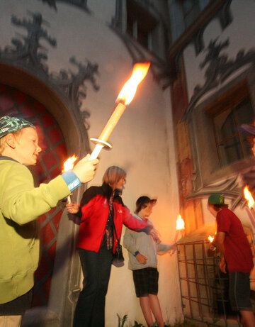 Das Bild zeigt eine lebendige Szene einer Gruppe von Menschen, die sich vor einem Gebäude versammeln. Das Gebäude, gekennzeichnet durch einen großen Bogen, dient als Kulisse für die Gruppe. Die Menschen, ein Gemisch aus Erwachsenen und Kindern, sind mit dem Aktes des Kerzenanzündens beschäftigt. Die Kerzen, von der Gruppe erhoben, brennen, und werfen ein warmes Schein auf die Szene. Die Gruppe ist vielfältig, mit Individuen unterschiedlichen Alters und Geschlechts. Einige von ihnen halten Fackeln, was zur feierlichen Stimmung beiträgt. Das Gesamtbild ist eine Feier und Einheit.