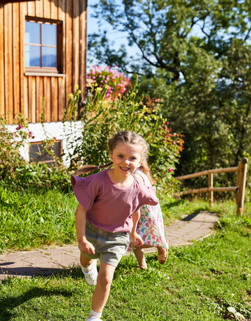Das Bild zeigt ein junges Mädchen, das in einem pinken Hemd und grauen Shorts spazieren geht. Sie lächelt und scheint ihre Zeit im Freien zu genießen. Der Weg, auf dem sie spaziert, ist von einer Holzlattenwand an der einen Seite und einer Vielzahl von Blumen an der anderen Seite gesäumt. Das Haus im Hintergrund ist aus Holz, hat ein rotes Dach und sieht aus, als wäre es an einem klaren und blauen Himmel an einem sonnigen Tag. Die Gesamtszene wirkt friedlich und idyllisch, und der fröhliche Ausdruck des Mädchens verleiht dem Bild ein Gefühl von Wärme und Glück.