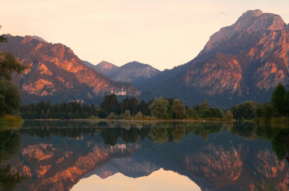 Das Bild zeigt eine friedliche Landschaft, die von einem majestätischen Gebirge beherrscht wird, das sich bis zum Horizont erstreckt. Die Berge, mit ihren schroffen Gipfeln und üppig grünen Wäldern, sind von dem warmen Schein der untergehenden Sonne überflutet. Der Himmel, in Tönen von Orange und Pink getaucht, wirft ein Reflexion auf den ruhigen Forggensee, der im Vordergrund liegt. Der See, mit seinen stillen Wassern, spiegelt die atemberaubende Berglandschaft und das Schloss Neuschwanstein wider, das sich auf einer Anhöhe vor dem Bergpanorama befindet.