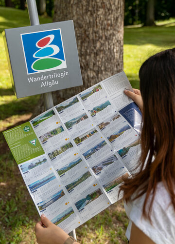 Das Bild zeigt eine Frau, die einen Flyer liest. Man sieht die Frau nur von hinten. Rings um sie herum wachsen Bäume und Gras. Die Sonne scheint. Auf einem Metallstab vor der Frau ist ein grauer Würfel befestigt. Unter einem Symbol ist „Wandertrilogie Allgäu“ zu lesen. 
