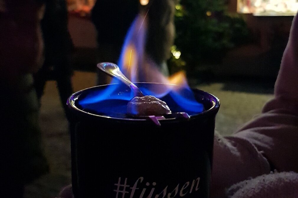 Das Bild zeigt eine Tasse. Auf dieser ist #füssen zu lesen. Auf der Tasse lodert eine kleine blaue Flamme. Ein Löffel liegt auf der Tasse. Im Hintergrund sin beleuchtete Weihnachtsmarktbuden zu sehen. Es ist Abend. 