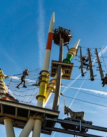 Das Bild zeigt eine aufregende Szene in einem Vergnügungspark. Der Hauptfokus liegt auf einer großen, bunten Anlage, die einem Klettergerüst ähnelt. Diese Anlage besteht aus verschiedenen hölzernen Pfosten und Plattformen, die durch eine Vielzahl von Seilen und Drähten verbunden sind.  Mehrere Personen sind zu sehen, die mit dieser Anlage interagieren, einige von ihnen klettern die Polster hinauf, während andere von den Plattformen schwingen. Die Anlage ist nicht nur ein Spielplatz, sondern auch ein Ort für Abenteuer.  Im Vordergrund befindet sich eine kleine, hölzerne Plattform mit einem Geländer. Diese Plattform ziert eine Statue eines Ziegen, die der Szene eine spielerische Note verleiht.  Der Himmel über ist ein klarer Blau, gesprenkelt mit weißen Wolken, und bietet eine atemberaubende Kulisse für die lebendige Anlage. Das Bild ist eine lebhafte Darstellung von Spaß und Abenteuer in einem Vergnügungspark.
