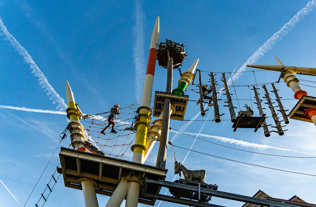 Das Bild zeigt eine aufregende Szene in einem Vergnügungspark. Der Hauptfokus liegt auf einer großen, bunten Anlage, die einem Klettergerüst ähnelt. Diese Anlage besteht aus verschiedenen hölzernen Pfosten und Plattformen, die durch eine Vielzahl von Seilen und Drähten verbunden sind.  Mehrere Personen sind zu sehen, die mit dieser Anlage interagieren, einige von ihnen klettern die Polster hinauf, während andere von den Plattformen schwingen. Die Anlage ist nicht nur ein Spielplatz, sondern auch ein Ort für Abenteuer.  Im Vordergrund befindet sich eine kleine, hölzerne Plattform mit einem Geländer. Diese Plattform ziert eine Statue eines Ziegen, die der Szene eine spielerische Note verleiht.  Der Himmel über ist ein klarer Blau, gesprenkelt mit weißen Wolken, und bietet eine atemberaubende Kulisse für die lebendige Anlage. Das Bild ist eine lebhafte Darstellung von Spaß und Abenteuer in einem Vergnügungspark.